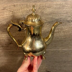 Può includere: Una teiera in metallo dorato con un design ornamentale e tre gambe. La teiera ha un coperchio con un pomo in cima.