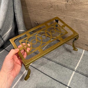 May include: A gold-colored metal trivet with a rectangular frame and decorative circular and geometric cutouts. The trivet has four curved legs and is designed to protect surfaces from hot cookware.