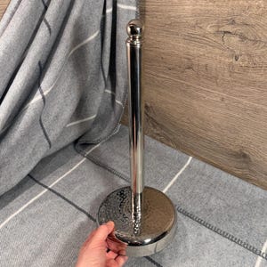 May include: A polished silver-tone metal kitchen roll holder featuring a tall, cylindrical vertical rod topped with a small sphere and a circular base with a textured, hammered finish. The stand is designed to hold a standard roll of kitchen paper.