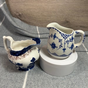 May include: Two blue and white ceramic creamer pitchers with floral designs. The pitcher on the left has a more intricate design with a red accent. The pitcher on the right has a simpler design with a white background.