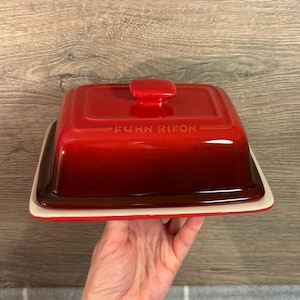 May include: A red ceramic butter dish with a lid and a white base. The lid has a square shape with a small handle and the text "KUHN RIKON". The dish is held by a person.