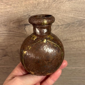 以下が含まれることがあります： 金色のアクセントが付いた、小さく丸い茶色の金属製の壺。壺は首が細く、底が広い。