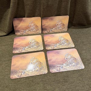 以下が含まれることがあります： 6枚の長方形のコースターセット。それぞれに、夕焼け色の暖かい空を背景に、背の高い乾いた草の中で休むヒョウの画像がプリントされています。コースターは角が丸く、滑らかで光沢のある仕上がりで、すべて同じ野生動物のシーンが描かれています。