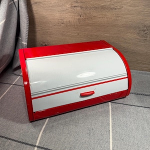May include: A rectangular bread bin with a vibrant red frame and a sliding roll-top door in a light grey finish. The door features thin horizontal grey stripes and a small, centred red handle for opening and closing the storage container.