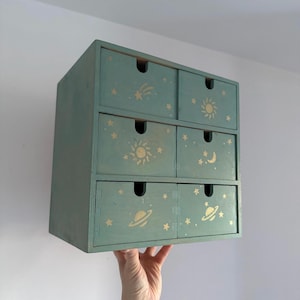 Könnte beinhalten: Eine kleine, teal-farbene Holzaufbewahrungstruhe mit sechs Schubladen. Jede Schublade ist mit goldenen Himmelsdesigns verziert, darunter Sterne, Sonnen, Monde und Planeten. Die Truhe wird vor einer weißen Wand gehalten.