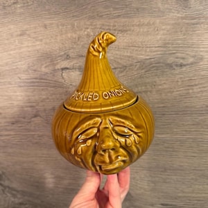 May include: A glossy mustard-yellow ceramic jar shaped like an onion with a lid. The lid features a pointed top and the text PICKLED ONIONS embossed in white. The base depicts a sculpted face with closed eyes and tear-like details on the cheeks.