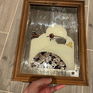 May include: Vintage framed Vogue magazine cover art. The artwork features a woman in a white hat and coat, with a brown car in the background. The frame is made of wood.