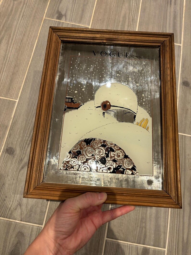 May include: Vintage framed Vogue magazine cover art. The artwork features a woman in a white hat and coat, with a brown car in the background. The frame is made of wood.