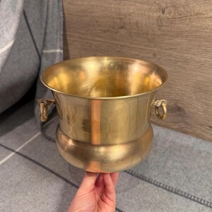 Puede incluir: Una jardinera de latón vintage con base redondeada y dos asas ornamentadas. La jardinera tiene una amplia abertura y un acabado pulido, que refleja la luz. La jardinera está sujeta por la mano de una persona.