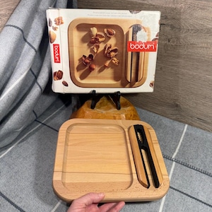 May include: A wooden nutcracker set by Bodum. The set includes a rectangular wooden tray with a recessed area and a nutcracker tool. The box shows the tray with nuts and shells. The Bodum logo is visible on the box.