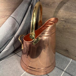 Può includere: Un secchio per carbone in rame lucido con un'ampia apertura inclinata e una robusta maniglia curva in ottone fissata con rivetti decorativi. Il contenitore presenta una maniglia in ottone più piccola sul lato e una base liscia e arrotondata.