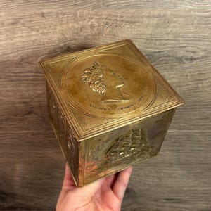 Puede incluir: Una caja de latón vintage con un relieve detallado de un perfil y un barco. La caja es cuadrada con una tapa con bisagras. La tapa presenta un relieve circular de un perfil con la inscripción "ELIZABETH II REGINA JANE". Los lados de la caja tienen un relieve de barco.