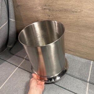 Puede incluir: Cubeta de hielo de acero inoxidable con base redondeada. La cubeta tiene forma cilíndrica con una parte superior ligeramente inclinada. La superficie metálica refleja la luz. Diseñada para enfriar bebidas. Aproximadamente 15 cm de altura.