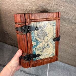 May include: A wooden box shaped like a book with a world map design on the front. The box has metal hinges and clasps.
