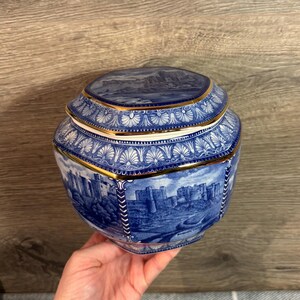 Pode incluir: Um pote de cerâmica azul e branco com acabamento dourado. O pote é octogonal e apresenta uma imagem detalhada de um castelo na frente. A tampa também é decorada com um design azul e branco.