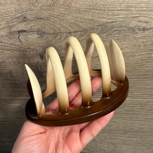 May include: A vintage, two-tone brown and cream-colored toast rack. The rack has a curved design with vertical slots for holding toast. The base is a darker brown, and the vertical supports transition from cream to brown.