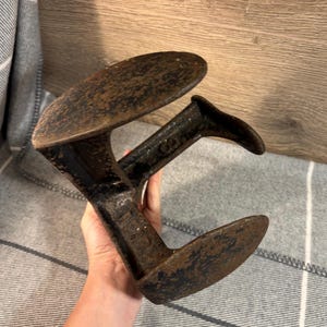 Puede incluir: Un estirador de zapatos vintage de hierro fundido con una pátina marrón oscura. El estirador tiene dos brazos que tienen la forma de la suela de un zapato. Los brazos están conectados por una barra de metal. El estirador está marcado con el número "62".