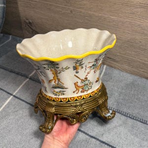 May include: A decorative ceramic vase with a scalloped rim and a yellow border. The vase features a detailed illustration of a hunting scene and sits on a gold-colored metal stand with ornate feet. The vase is held by a person's hand.