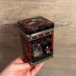 Könnte beinhalten: Eine rechteckige schwarze Blechdose mit asiatisch inspiriertem Design. Die Dose hat rote und goldene Akzente mit Illustrationen von Figuren und Kirschblüten. Der Deckel hat ein kreisförmiges Design mit Vögeln.