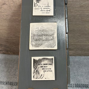 May include: A grey wooden box with three white tiles featuring inspirational quotes. The top tile says "Be a rainbow in someone else's cloud." - Maya Angelou. The middle tile says "Life isn't about finding yourself. Life is about creating yourself." The bottom tile says "Positive thinking evokes more energy, more initiative, more happiness."