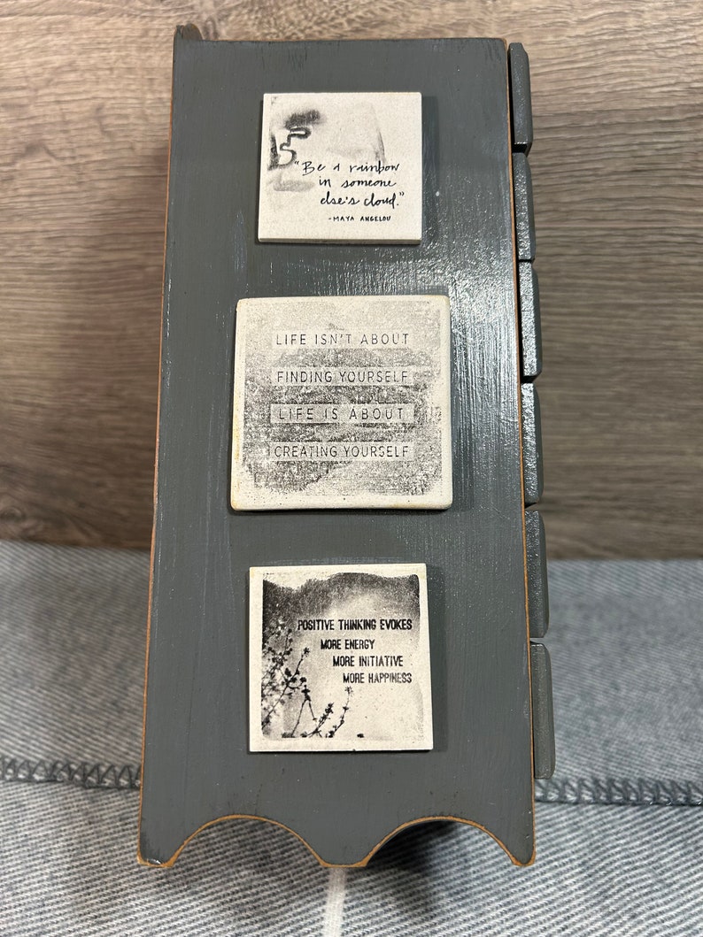 May include: A grey wooden box with three white tiles featuring inspirational quotes. The top tile says "Be a rainbow in someone else's cloud." - Maya Angelou. The middle tile says "Life isn't about finding yourself. Life is about creating yourself." The bottom tile says "Positive thinking evokes more energy, more initiative, more happiness."