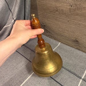 May include: A hand bell featuring a polished brass bell body with a rounded base and a turned wooden handle. The handle has a dark brown finish with decorative horizontal grooves and a small rounded knob at the very top of the grip.