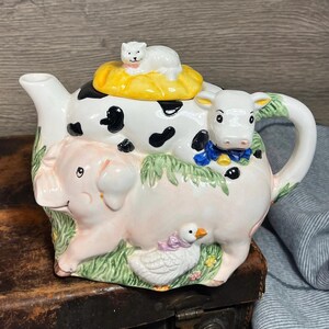 Op de afbeelding: Een grillige keramische theepot met boerderijdieren. De theepot is wit met zwarte vlekken, een roze varken, een witte koe met een blauwe strik en een witte kat bovenop. De theepot heeft een wit handvat en tuit.
