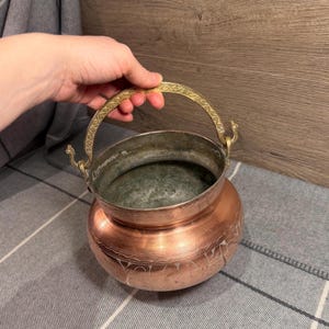 Puede incluir: Un caldero de cobre con asa de latón. El caldero tiene diseños grabados alrededor de la base y un interior deslustrado. El asa es arqueada y está decorada con un patrón repetitivo.