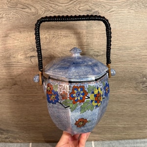 Puede incluir: Una cubitera de cerámica azul y blanca con un asa trenzada negra. La cubitera presenta un diseño floral en rojo, naranja, amarillo y azul. La tapa tiene un pequeño pomo en la parte superior.