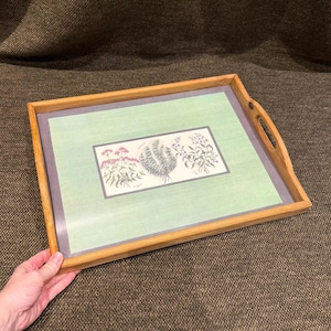 May include: A rectangular wooden serving tray with integrated side handles. The tray features a light green central panel with a grey border, containing a botanical illustration of three distinct herb clusters with pink and green foliage.