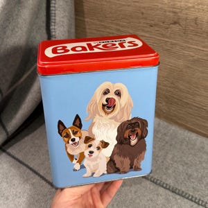 May include: A rectangular tin with a red lid and a light blue body. The lid has the word "Bakers" in white. The body features illustrations of four different dog breeds. The tin is designed for pet food storage.