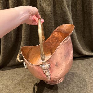 May include: A polished copper coal scuttle featuring a wide, open-top design with a curved brass handle. The vessel has a rounded body, a flared rim, and decorative brass attachments where the handle connects to the main copper structure.