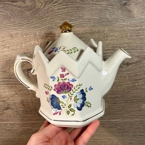 Può includere: Teiera in ceramica bianca con coperchio e manico dorati. La teiera presenta un motivo floreale con fiori viola e blu, foglie verdi e finiture dorate. Design angolare unico.