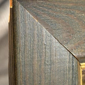 Puede incluir: Primer plano de la esquina de un marco de imagen de madera. El marco es de color marr&oacute;n oscuro con un patr&oacute;n de veta de madera. El marco tiene un aspecto desgastado con un lavado gris claro sobre la madera.