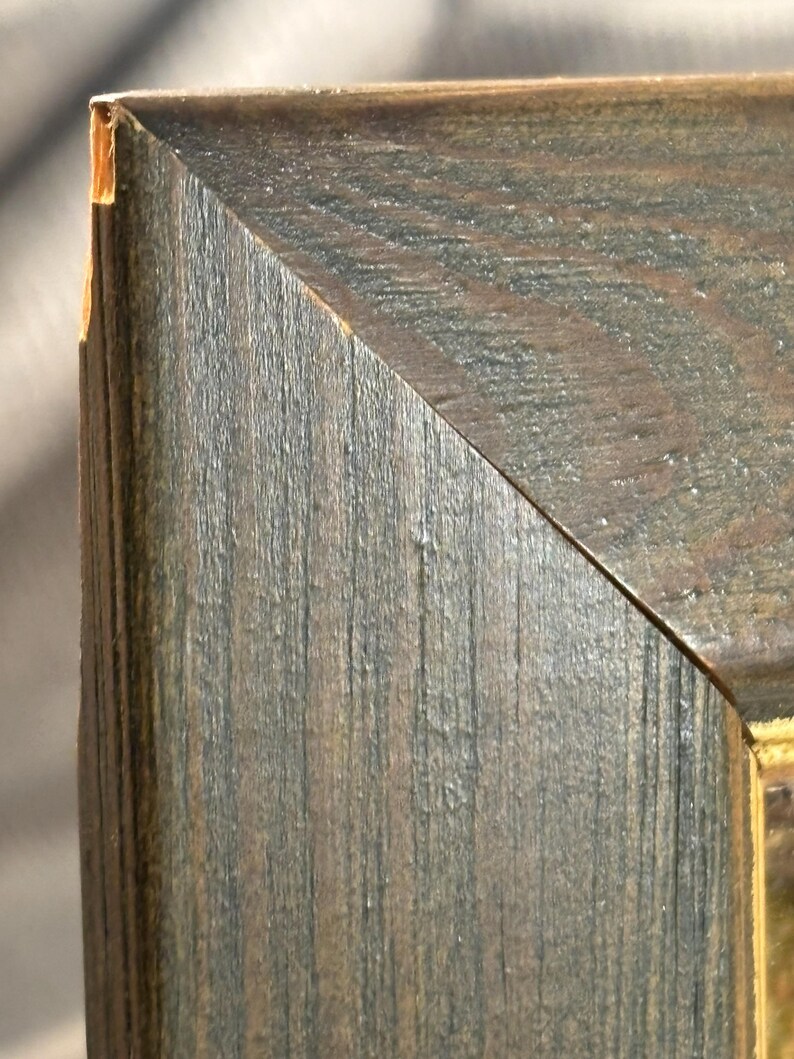 Puede incluir: Primer plano de la esquina de un marco de imagen de madera. El marco es de color marr&oacute;n oscuro con un patr&oacute;n de veta de madera. El marco tiene un aspecto desgastado con un lavado gris claro sobre la madera.