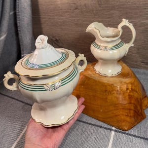 May include: A vintage cream-colored sugar bowl and creamer set with green and gold trim. The sugar bowl has a lid with a floral design. Both pieces have a delicate, scalloped edge.