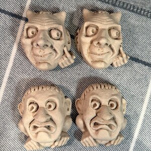 May include: Four small, off-white, sculpted faces with exaggerated features. Two faces have a mischievous expression, with one finger raised. The other two faces have a frowning expression. The faces are arranged on a blue and white striped fabric.