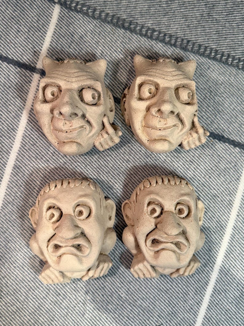 May include: Four small, off-white, sculpted faces with exaggerated features. Two faces have a mischievous expression, with one finger raised. The other two faces have a frowning expression. The faces are arranged on a blue and white striped fabric.