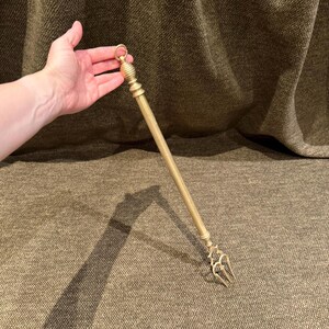 May include: A long, slender brass fireplace poker featuring a smooth, cylindrical shaft, a decorative ribbed handle topped with a small circular hanging ring, and a three-pronged, curved metal tip designed for manipulating logs and embers.