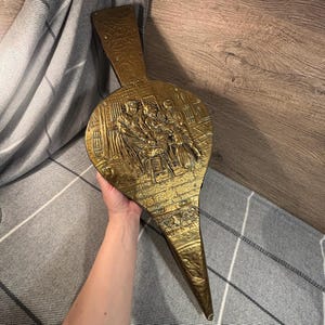 Könnte beinhalten: Ein antiker Messingbalg mit einer geprägten Szene von Figuren in einem Raum. Der Balg hat einen langen Griff und eine spitze Düse. Das Messing hat einen warmen, goldenen Farbton, und das Design ist detailliert und verziert.