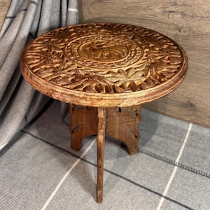 May include: A round, wooden table with intricate carvings. The table has a three-legged base and a top that is decorated with a floral pattern.