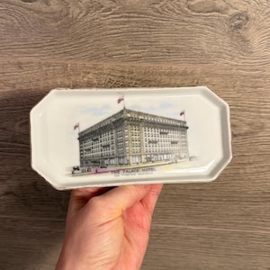 May include: A rectangular white ceramic dish with a gold trim. The dish features a detailed illustration of The Palace Hotel in San Francisco, California, with text below the image. The dish is held by a person.