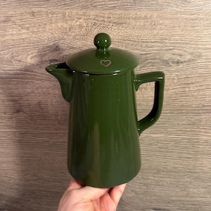 May include: A dark green enamel coffee pot with a lid and handle. The lid has a small heart-shaped detail. The pot is held by a person's hand.