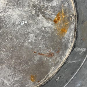 Op de afbeelding: De binnenkant van een ronde metalen bak met een verweerd, gegalvaniseerd grijs oppervlak met zichtbare tekenen van oxidatie. De bodem vertoont vlekken van oranje roest en een gevlekte grijze verkleuring, met een duidelijke naad.