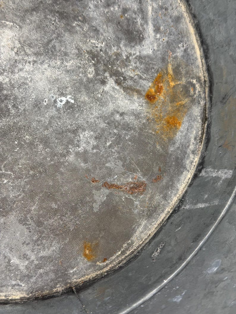 Op de afbeelding: De binnenkant van een ronde metalen bak met een verweerd, gegalvaniseerd grijs oppervlak met zichtbare tekenen van oxidatie. De bodem vertoont vlekken van oranje roest en een gevlekte grijze verkleuring, met een duidelijke naad.