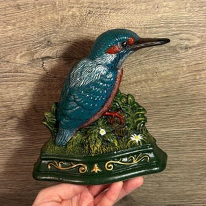 Könnte beinhalten: Ein blauer und grüner Türstopper aus Gusseisen in Form eines Eisvogels, der auf einem Bett aus grünen Blättern mit weißen Blüten sitzt. Der Türstopper hat eine grüne Basis mit goldenen Akzenten.