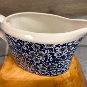May include: A white ceramic gravy boat with a blue and white floral pattern. The boat has a handle and a spout.
