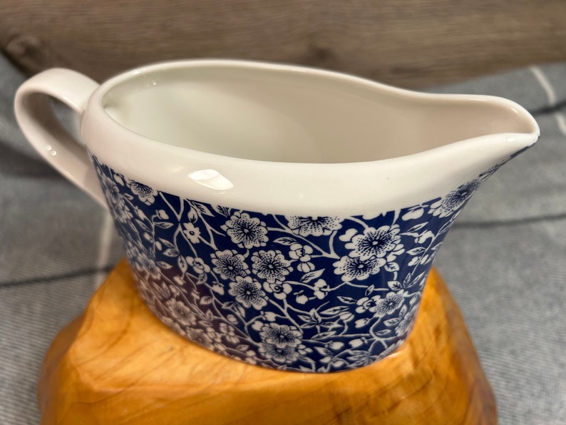 May include: A white ceramic gravy boat with a blue and white floral pattern. The boat has a handle and a spout.