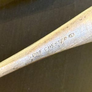 Op de afbeelding: Close-up van een zilverkleurig metalen object met de inscriptie "LOWE CHESTER". Het object lijkt deel uit te maken van een groter voorwerp, zoals een keukengerei of gereedschap. Het oppervlak vertoont slijtage.