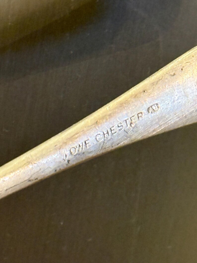 Op de afbeelding: Close-up van een zilverkleurig metalen object met de inscriptie "LOWE CHESTER". Het object lijkt deel uit te maken van een groter voorwerp, zoals een keukengerei of gereedschap. Het oppervlak vertoont slijtage.